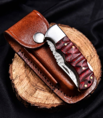 Damascus steel folding knife with sculpted red wood handle resting half-open on its leather belt pouch